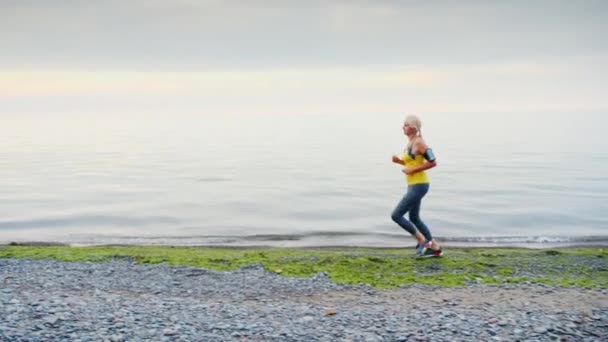 Een jonge vrouw jogs langs de oever van de zee of een meer, herfst, ochtend — Stockvideo