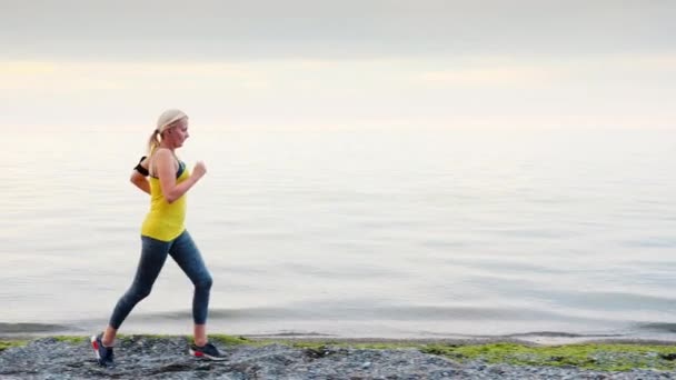 Una donna con una maglietta gialla corre lungo la riva di un lago o di un mare — Video Stock