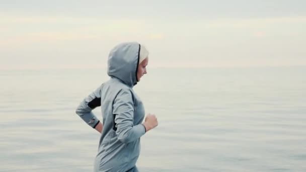 Correndo com o tempo frio. Uma mulher de fato desportivo e um capuz a correr à volta do mar ou do lago. Steadicam tiro — Vídeo de Stock