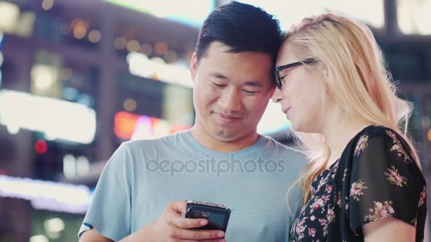 Una joven pareja multiétnica utiliza un teléfono inteligente. Ciudad nocturna sobre un fondo de luces borrosas — Vídeos de Stock