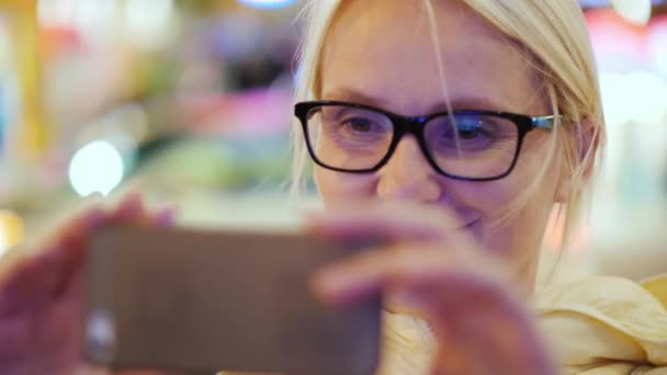 Una mujer turística alegre fotografía con la ayuda de teléfonos inteligentes. Primer plano en una calle concurrida por la noche, luces publicitarias están ardiendo, el fondo está borroso — Vídeos de Stock