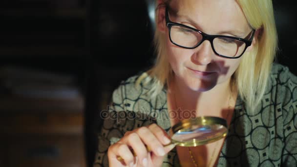 En kvinna i glasögon ser något genom ett förstoringsglas. Noggrann undersökning — Stockvideo