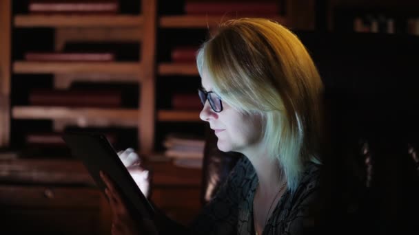 Vista lateral: Uma mulher de óculos trabalha com um tablet no fundo de uma prateleira com livros. Biblioteca conceitual, trabalho ou estudo até tarde — Vídeo de Stock