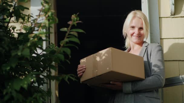 En kvinna med ett paket står på tröskeln till sitt hus. Ser på kameran, leende. Leverans av paket till dörren — Stockvideo