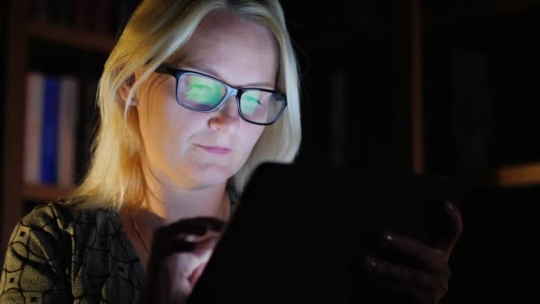 A középkorú nő dolgozik késő estig, élvezi a tabletta. A képernyőn a tabletta tükröződik a szemüvegét — Stock videók