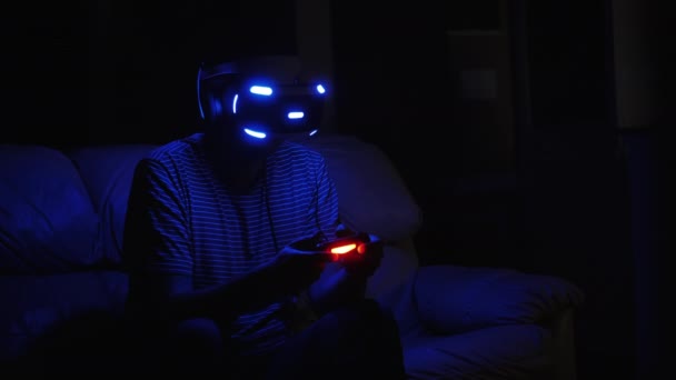 Un hombre juega juegos de ordenador en un casco de realidad virtual. El casco y el joystick están encendidos. Está oscuro en la habitación. — Vídeo de stock