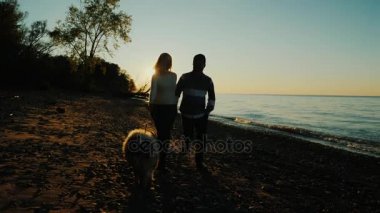 Çok ırklı çift ile deniz ya da göl, bir köpeğe günbatımında yürüme