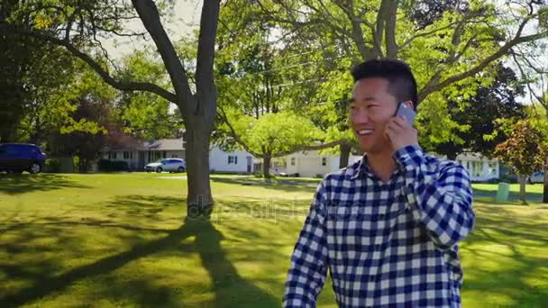 Молодой азиат идет по улице и разговаривает по телефону. Типичный пригородный город США — стоковое видео