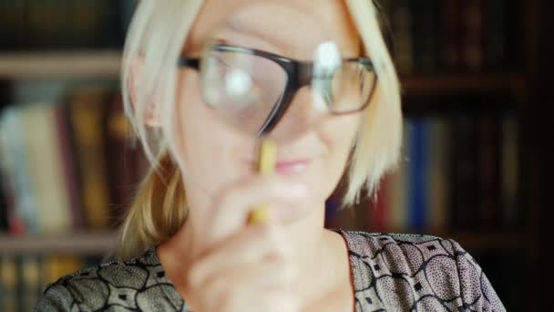 女性は、虫眼鏡を通してカメラに見えます。本と棚の背景のライブラリに立っています。 — ストック動画