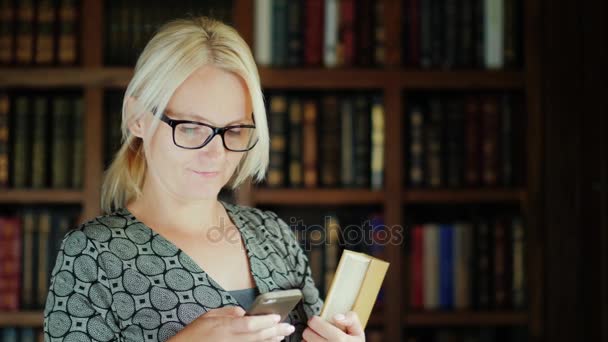 Μια γυναίκα με τα γυαλιά χρησιμοποιεί ένα smartphone στη βιβλιοθήκη, κρατά τα βιβλία στα χέρια της. Φόντο από τα ράφια με τα βιβλία — Αρχείο Βίντεο