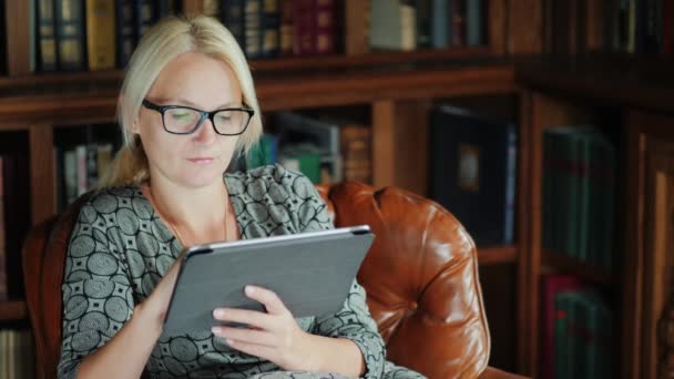 Bir iş kadını tablet kullanma bir lüks deri sandalyede oturuyor. Rafları kitaplarla arka planı — Stok video