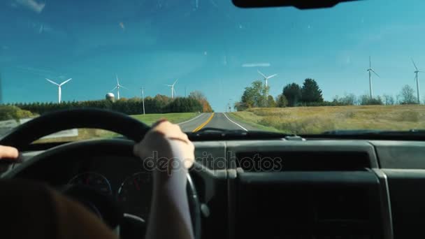 O motorista ao volante do carro vai em uma estrada rural. Nos lados há muitas turbinas eólicas. Ecoturismo e energia limpa — Vídeo de Stock