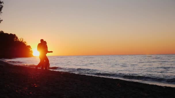 Ένα νεαρό ζευγάρι παίζει με ένα σκυλί στην όχθη της λίμνης. Στο ηλιοβασίλεμα, βίντεο αργής κίνησης — Αρχείο Βίντεο