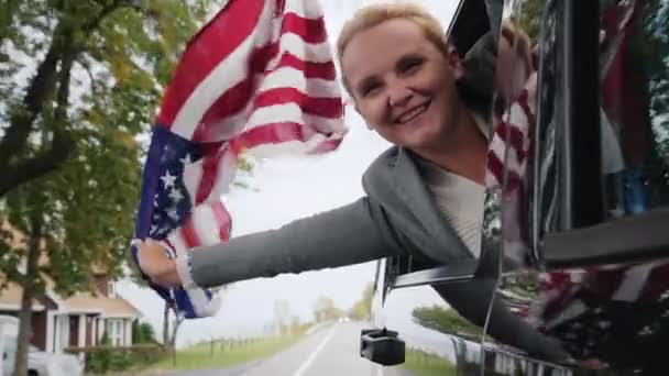 Een vrouw met een grote Amerikaanse vlag kijkt uit het raam van een reizende auto. Slow motion video — Stockvideo