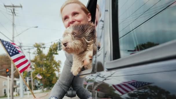 犬とアメリカの国旗を持つ女性は、車の窓から見えます。彼女はアメリカの郊外に沿って移動します。米国の旅行のコンセプト — ストック動画