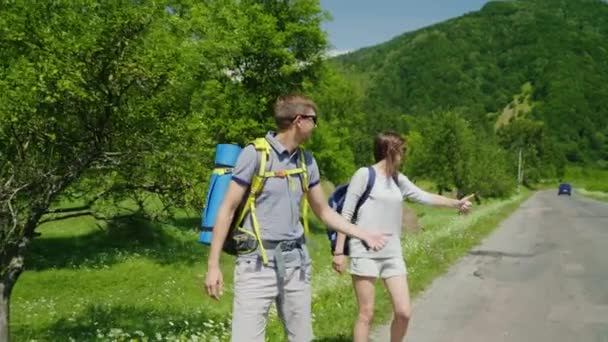 Пара туристов с рюкзаками едут автостопом по дороге, ловят проезжающую машину. Путешествия и приключения — стоковое видео