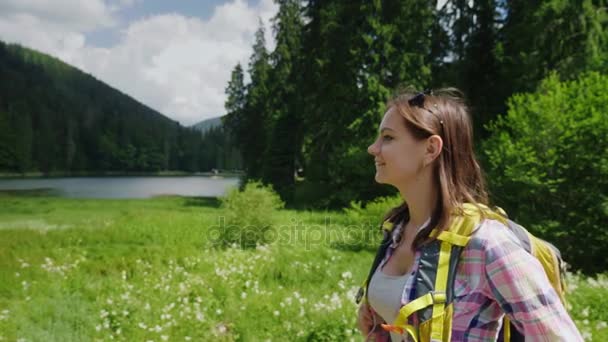 Een aantrekkelijke toeristische vrouw gaat naar een schilderachtige plaats op de achtergrond van weiden, bergen en hemel. Toerisme en actieve levensstijl — Stockvideo