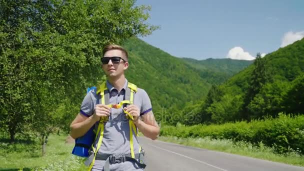 Atractivo turista hombre con una mochila caminando a lo largo de la carretera sobre un fondo de montañas verdes. Turismo, estilo de vida saludable y activo — Vídeos de Stock
