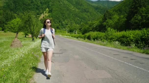 Yol boyunca yeşil dağların bir arka plan üzerinde yürüyen bir sırt çantası ile çekici kadın turist. Turizm, sağlıklı ve aktif bir şekilde yaşam — Stok video