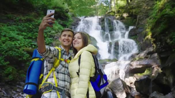 Et ungt par rejsende fotograferer sig selv på baggrund af et vandfald i bjergene. Kranskud – Stock-video