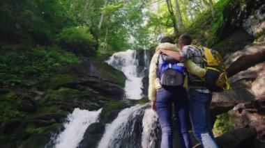 Arka görünümü: turist dağlarda şelale hayran sırt çantaları ile genç bir çift. Kaygan taşlar üzerinde durmak