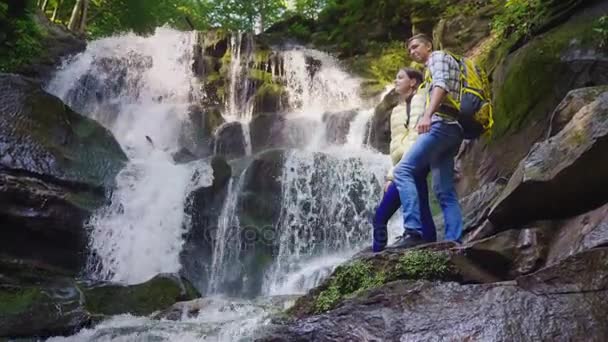 Молодая пара туристов с рюкзаками, любующихся водопадом в горах. Стоять на скользких камнях — стоковое видео