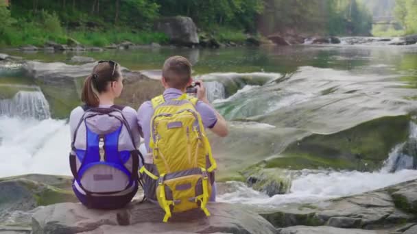 Egy fiatal pár, a turisták számára, gyönyörködni a hegyi folyó bevétel mozi-ból egy smartphone folyó ember — Stock videók