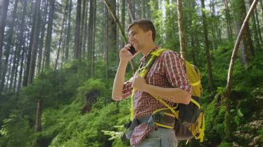 Genç adam ormanda telefonda konuşurken bir sırt çantası ile. Sabah güneş ışınlarının ağaçların dalları ile Parlatıcı