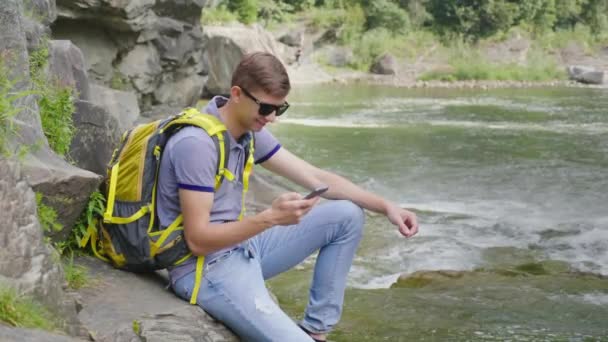 Молодой человек с желтым рюкзаком сидит у горной реки. Использует смартфон. Концепция туризма и технологий — стоковое видео