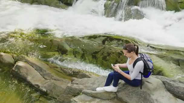 Vista superior: Una mujer joven se sienta en una roca sobre un fondo de un río de montaña. Utiliza un smartphone. Tecnología y concepto de viaje — Vídeos de Stock