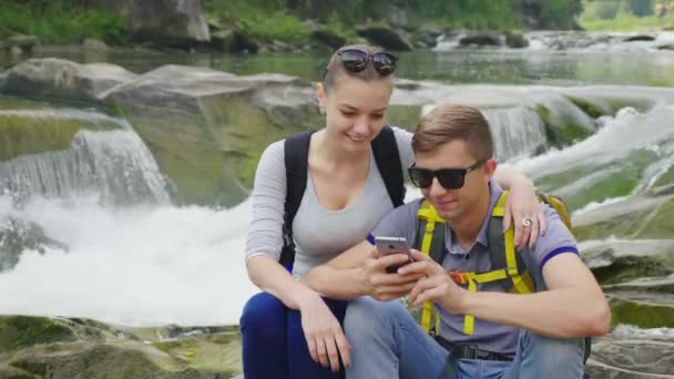 Genç bir çift bir engebeli dağ nehir çerçevede bir kaya üzerinde oturuyor. Bir akıllı telefon kullanıyorlar. Teknoloji ve seyahat kavramı — Stok video