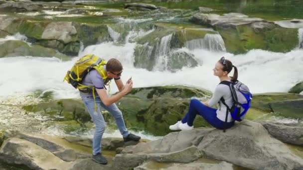 Un tipo toma fotos de su novia en un lugar pintoresco cerca de un río de montaña. Turismo y estilo de vida activo — Vídeo de stock