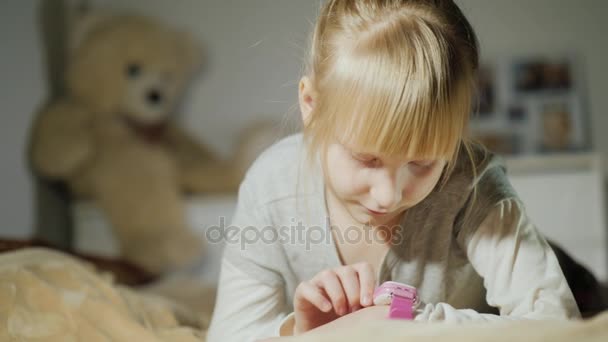 Une fille joue avec une horloge intelligente sur le bras. Allongé sur le lit dans la chambre — Video