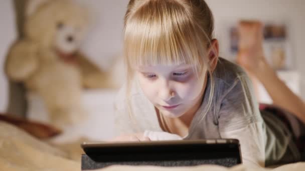 Маленька дівчинка лежить на ліжку у своїй спальні, використовуючи планшет — стокове відео