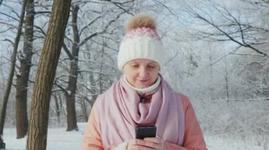 Genç kadın kış parkta yürüyor, smartphone sahiptir