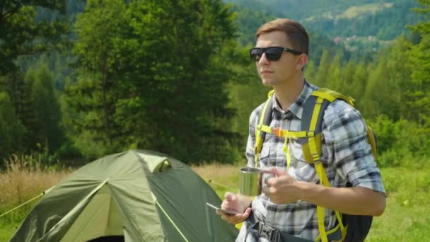 Aktive Lebensweise - ein junger Mann auf einem Campingplatz, genießt ein Telefon, trinkt Tee aus einem Becher — Stockvideo