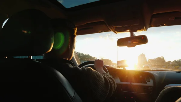 Un asiático conduce un coche a lo largo de la carretera, el sol poniente brilla en el parabrisas. Vista trasera — Foto de Stock