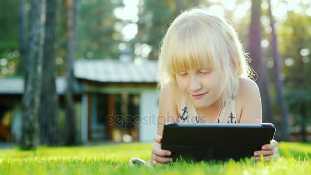 La chica está jugando en la tableta. El sol resalta maravillosamente su cabello rubio. Se encuentra en el césped cerca de la casa — Vídeos de Stock