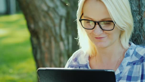 Привлекательная молодая женщина в очках пользуется планшетом. Сидит в парке возле дерева, красивый свет перед закатом. Вид сверху — стоковое видео