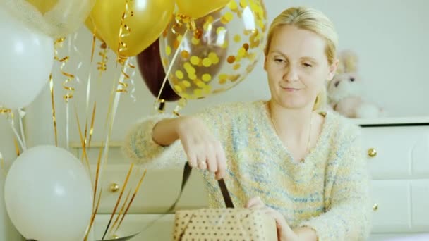 Maman enveloppe une boîte avec un cadeau dans du papier d'emballage. Assis près du lit dans la chambre d'enfants décorée de boules. Pour préparer une surprise pour l'anniversaire . — Video