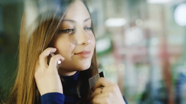 Uma jovem está ao pé da janela da loja. Tem um cartão de crédito na mão, a falar ao telefone. Grande conceito de compras — Vídeo de Stock