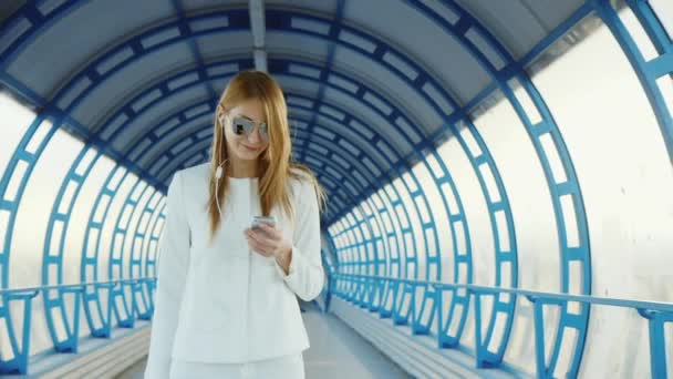Стильная деловая женщина в белом костюме проходит через туннель или переход от стекла. Счастливая сумка на колёсах, с мобильным телефоном — стоковое видео