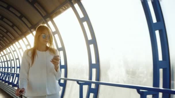 一个成功和有目的的女人走过玻璃隧道, 使用智能手机。夕阳照耀着她。企业女性理念 — 图库视频影像
