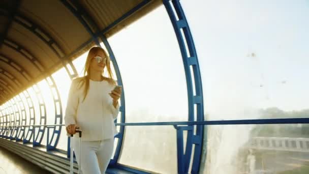 Een stijlvolle vrouw is reizen. In de stralen van de ondergaande zon gaat door een glazen tunnel met een tas op wielen, gebruikt een telefoon. — Stockvideo