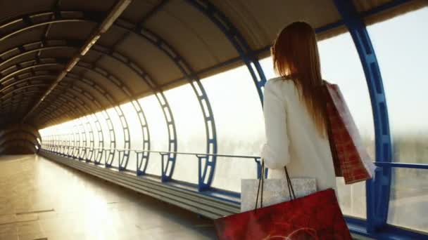 Weergave achterzijde: gelukkige vrouw met boodschappentassen. Glimlachen, wandelen langs een moderne glazen tunnel. Bezit van een smartphone — Stockvideo