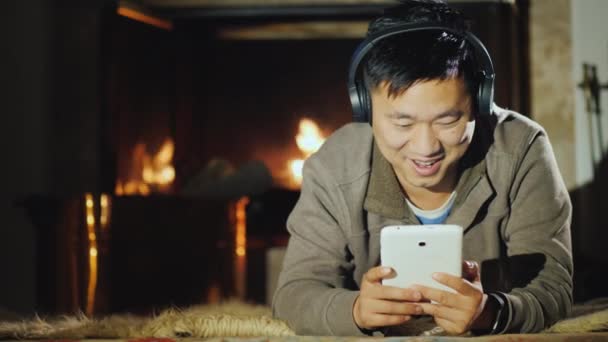 Happy Asian man resting at home by the fireplace, enjoys a tablet — Stock Video