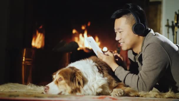 Un hombre asiático feliz yace junto a la chimenea con un perro. Utiliza una tableta digital — Vídeos de Stock