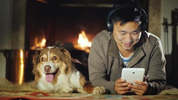 Een Chinese man rust bij de open haard, geniet van een tablet. Vervolgens is zijn hond — Stockvideo