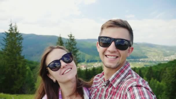 Um casal feliz em óculos de sol tira fotos de si mesmos em um lugar pitoresco no fundo das montanhas — Vídeo de Stock