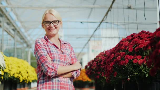 Young woman smiling, looking at camera. It stands on the background of flowers in a plant nursery — Stock Video